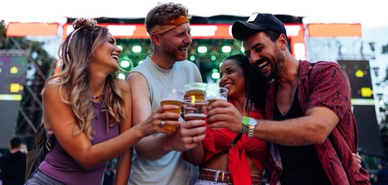 Cuatro asistentes brindando con cervezas frente al escenario de un festival